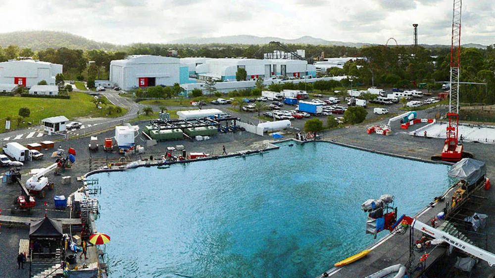 A large outdoor water tank surrounded by equipment, vehicles, and studio buildings, set in an industrial area with green hills and cloudy skies in the background.