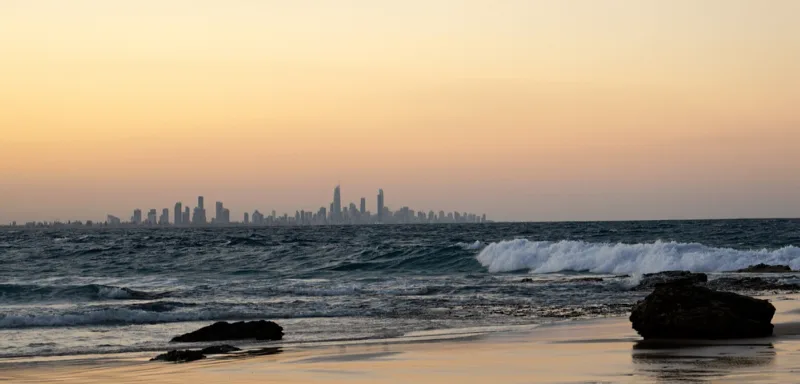Waves crash on a rocky beach at sunset with a distant city skyline visible on the horizon under a clear sky.