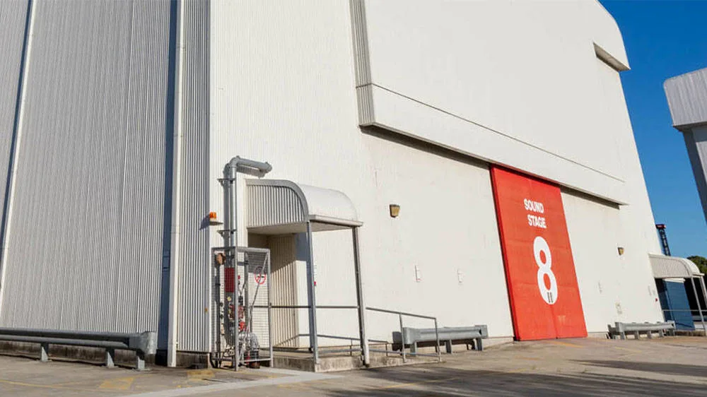 A large white industrial building with a red door labeled "Sound Stage 8" in white text. The area outside is paved, with metal railings and a covered entryway on the left.