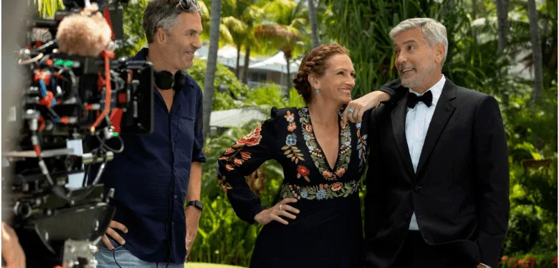 Three people are smiling outdoors on a film set: a man in casual clothes with a headset, a woman in a floral embroidered dress, and a man in a tuxedo, with a camera and greenery in the background.