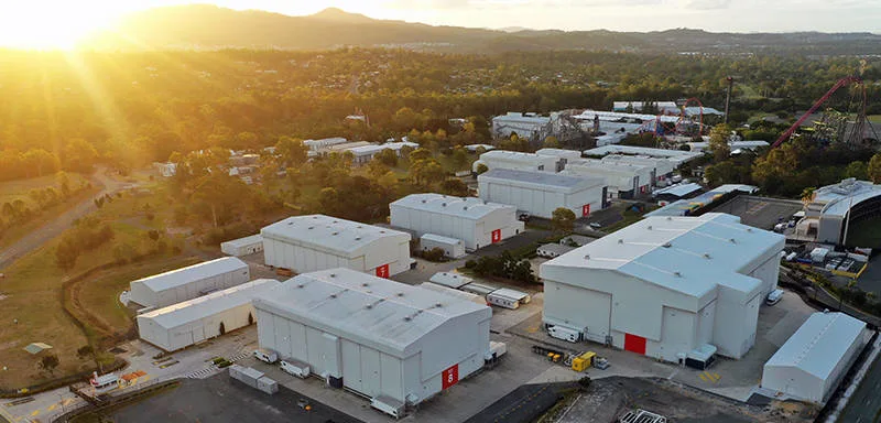 Aerial view of large white industrial buildings surrounded by trees at sunset, with mountains in the background and sunlight streaming in from the left side.
