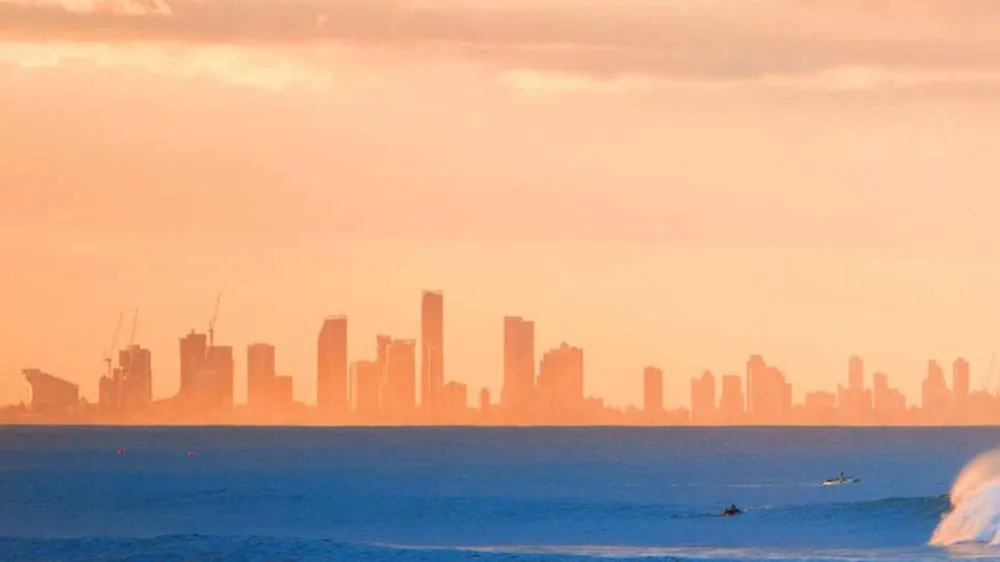 A city skyline with tall buildings is silhouetted against an orange sky at sunset, viewed from across the ocean with some waves and small boats visible in the foreground.