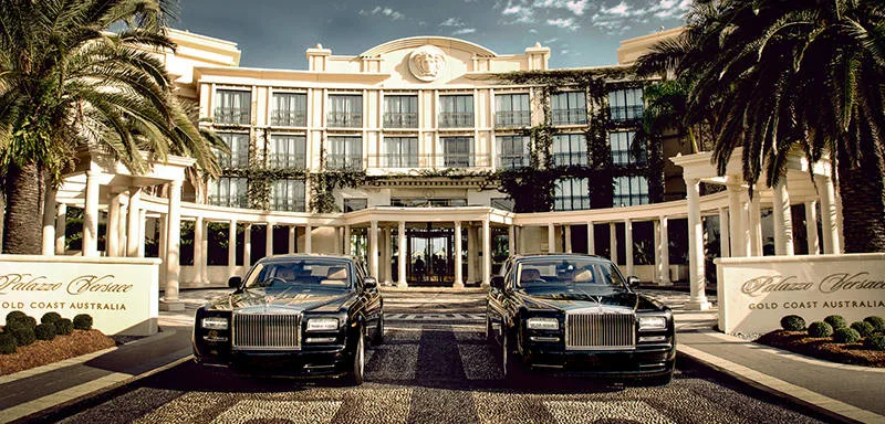 Luxury hotel entrance with grand columns and palm trees. Two black Rolls-Royce cars are parked symmetrically in front. The sign reads "Palazzo Versace Gold Coast Australia" in gold lettering.