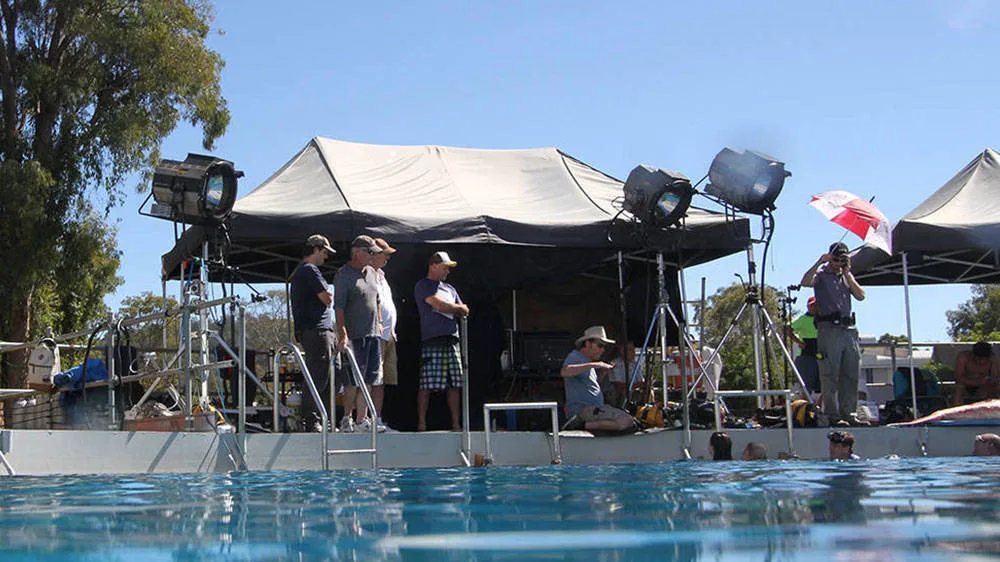 Film crew stands by a poolside set with large lights and equipment under tents, some people wearing hats and summer clothes, while the foreground shows the pool water under a clear blue sky.