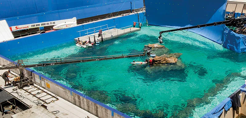 A film set with a large pool, artificial rocks, and crew working on platforms. Blue screens surround the water for visual effects, and cameras and equipment are visible around the set.