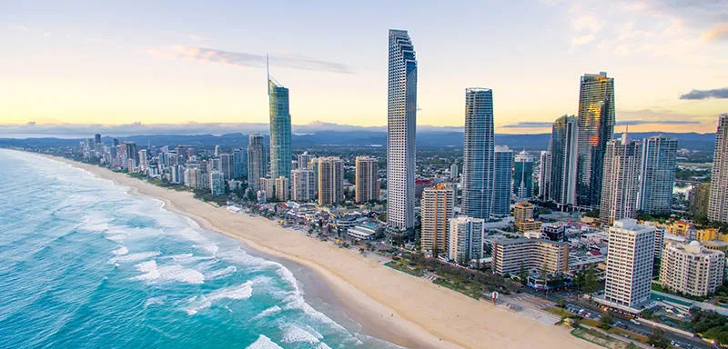 Aerial view of a coastal city with tall modern skyscrapers lining a sandy beach, bordered by blue ocean waves under a partly cloudy sky at sunset.