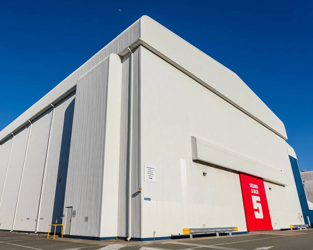Large, white industrial building with high walls and a prominent red door marked "STAGE 5" in white letters, under a clear blue sky.