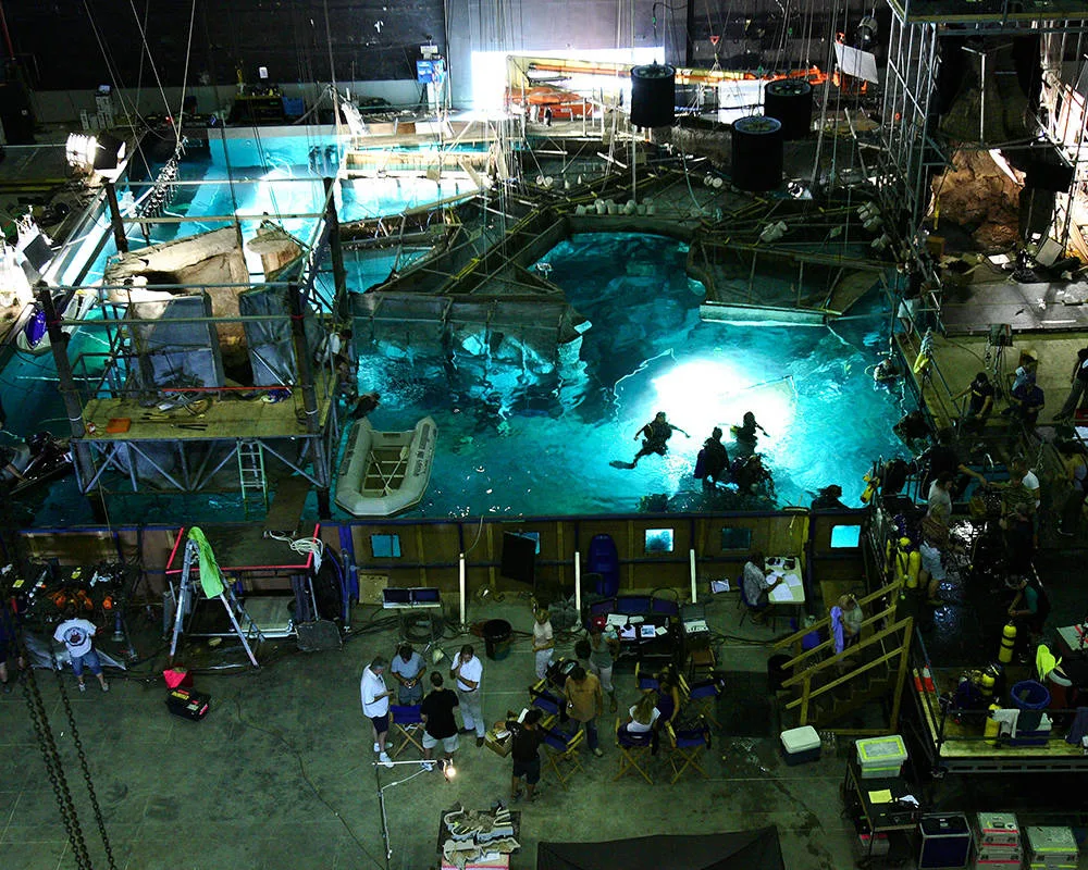 A behind-the-scenes view of a film set with crew members working around a large illuminated water tank, diving equipment, scaffolding, and various equipment scattered around the area.