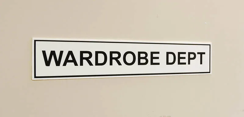 A white sign with black border reads "WARDROBE DEPT" in bold black letters, mounted on a beige wall.
