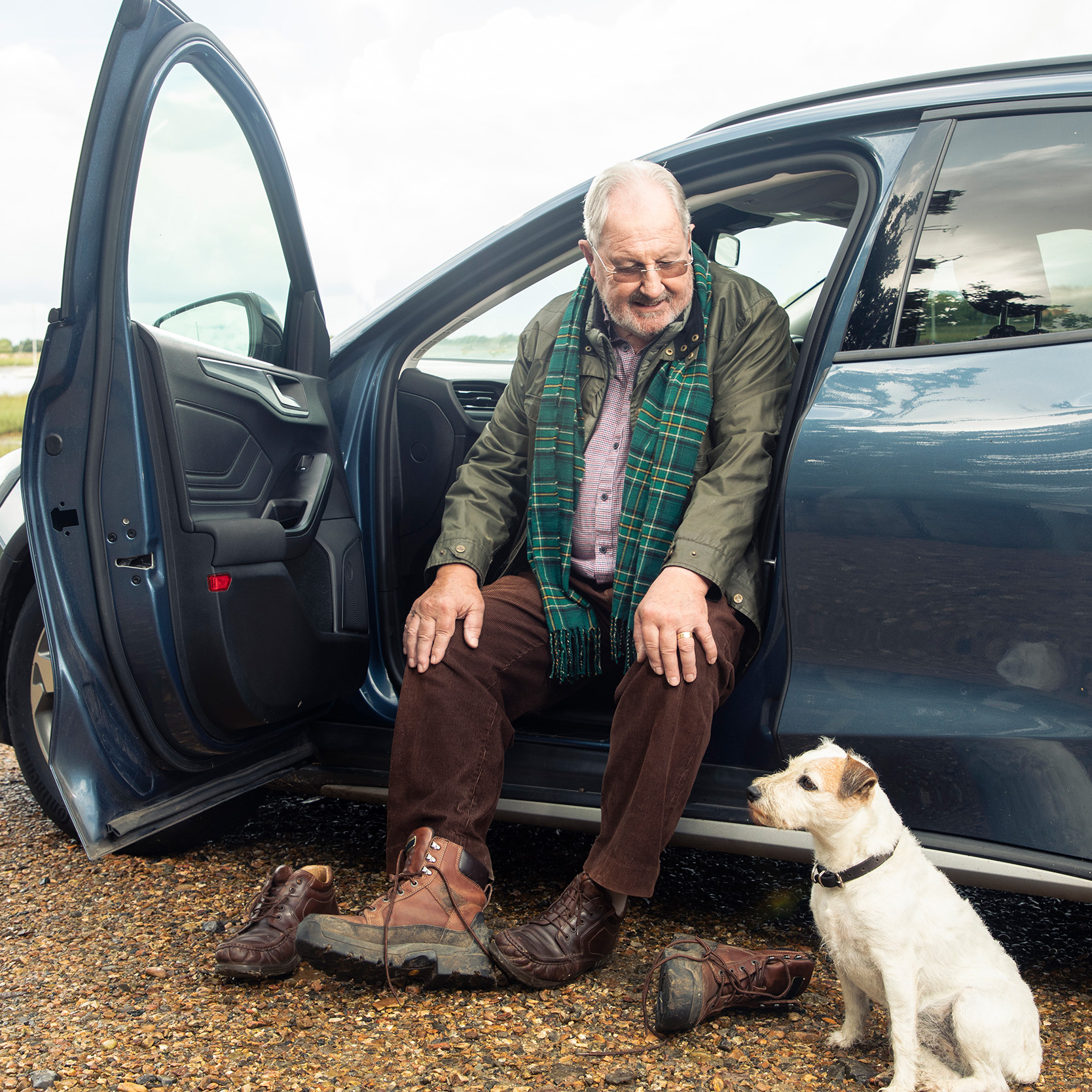 Man wearing olive jacket and tartan scarf sitting in open car door with small white terrier dog beside him.