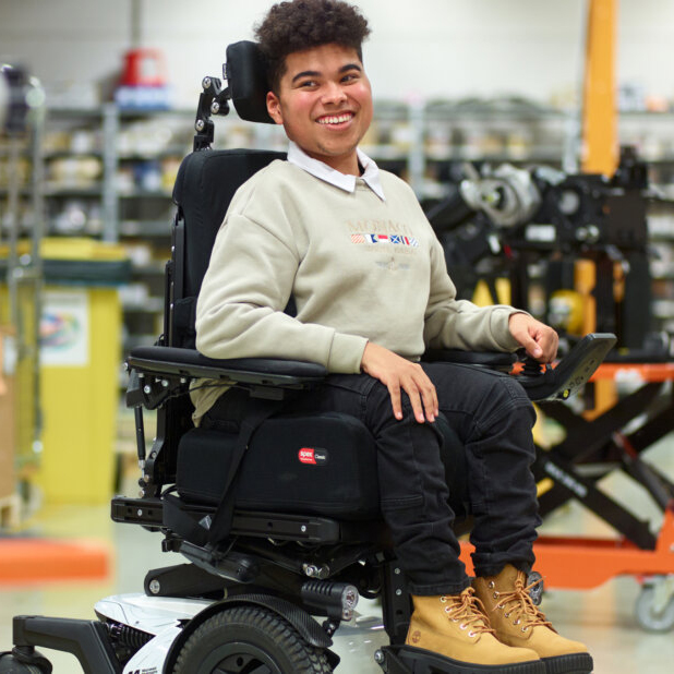 Young person smiling in a powered wheelchair wearing a beige jumper in a workshop with exercise equipment in the background.