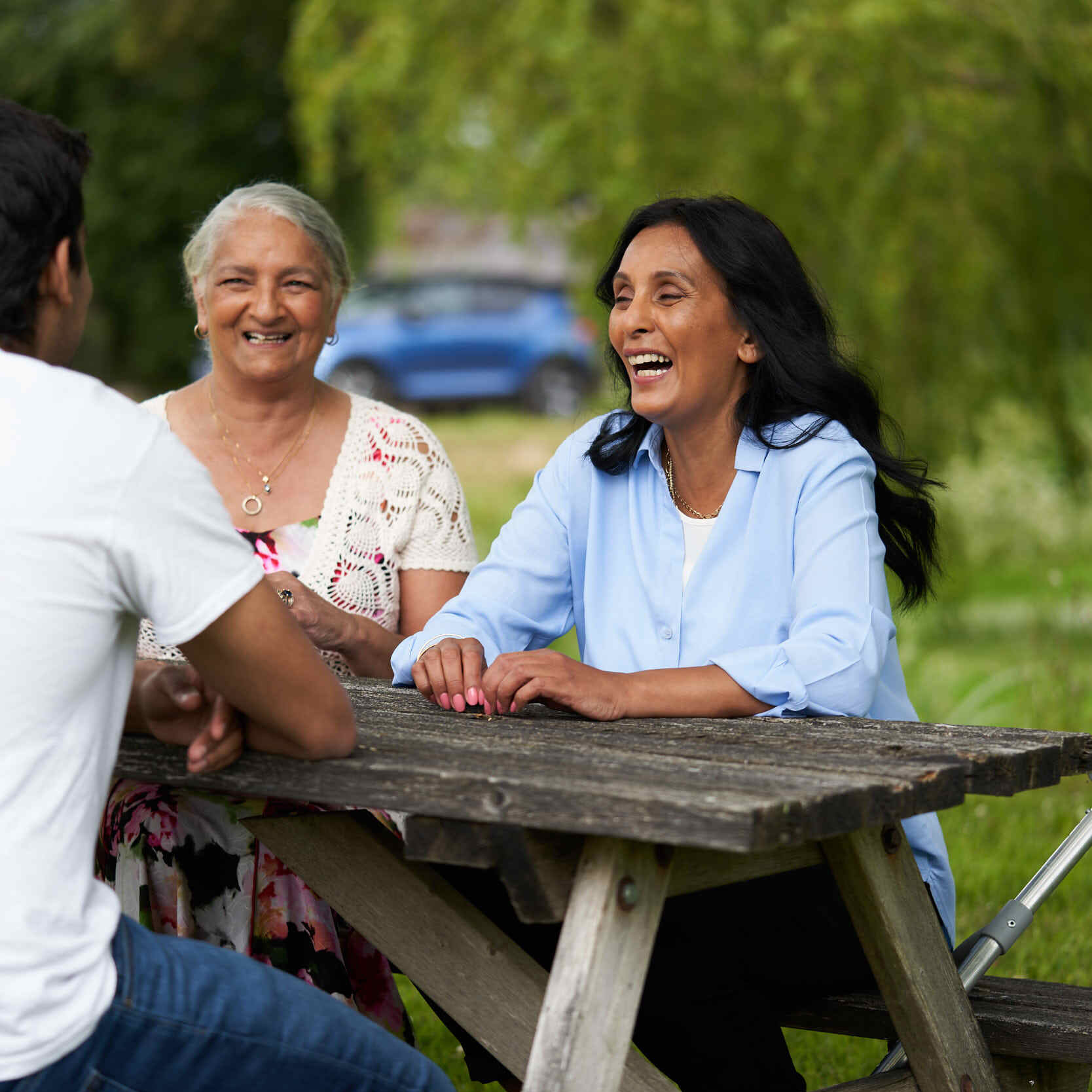 Three people laughing together at a wooden picnic table outdoors, with a blue car and lush green trees in the background.