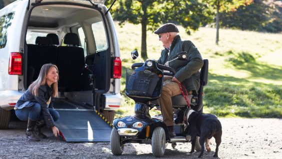 A woman lowering a rear ramp on a WAV, while a man on a mobility scooter talks to her