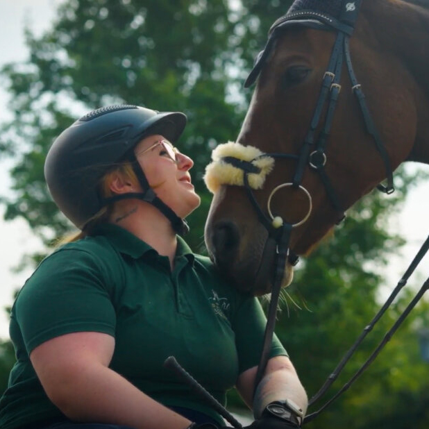 Woman in green polo shirt and riding helmet affectionately nuzzling a bay horse wearing a bridle outdoors.