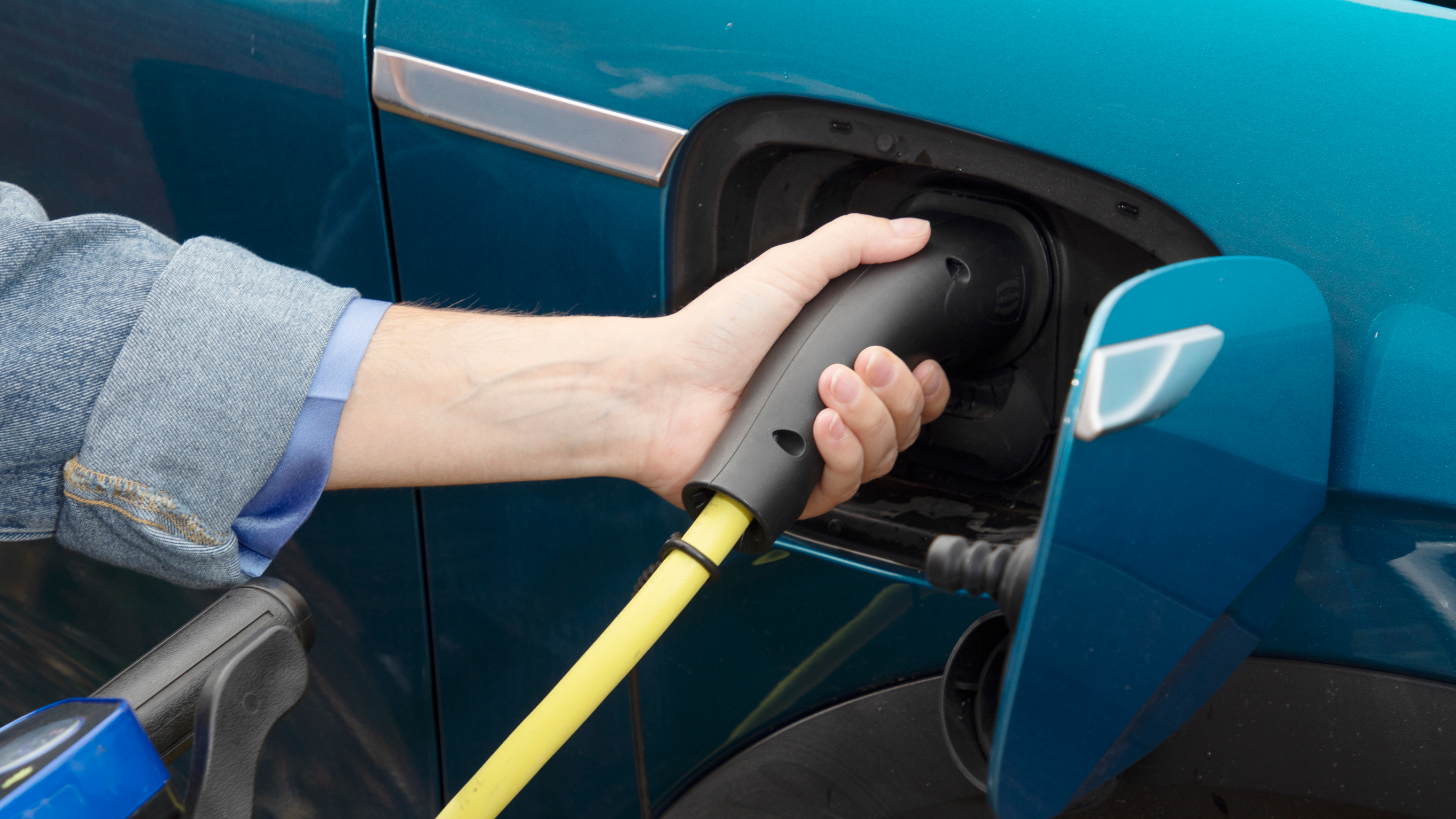 Close-up of a person’s hand plugging a yellow electric vehicle charging cable into a blue car.