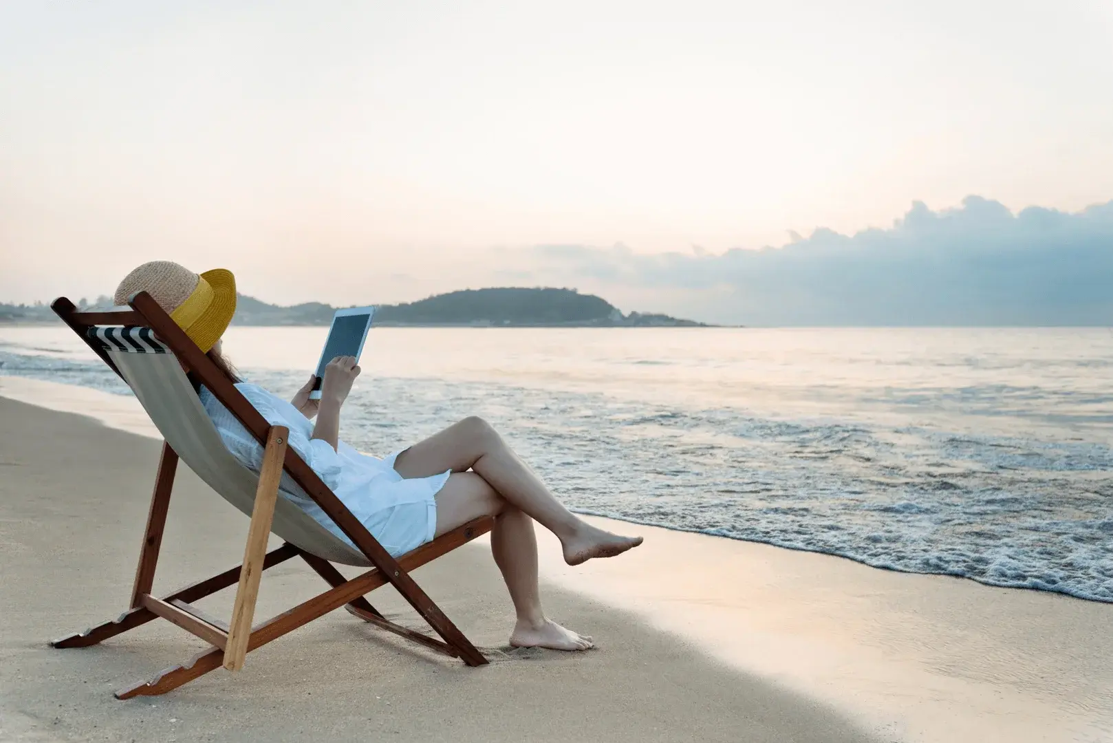 Eine Frau sitzt in einem Liegestuhl am Strand und schaut auf ihr iPad, während sie ihre freie Zeit genießt.