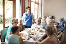 Foto: Im Pflegeheim sitzen ältere Damen am Mittagstisch und eine Pflegerin unterhält sich mit ihnen.