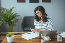 Foto: Eine Frau sitzt vor einem Laptop, Rechnungen liegen daneben. Sie hält Geld in den Händen.