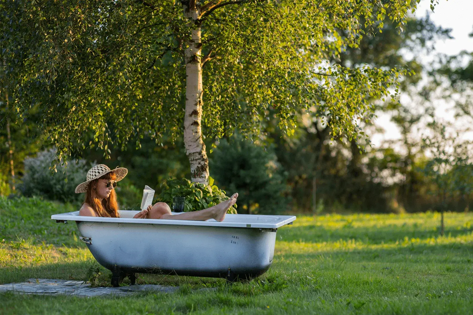 Eine Frau sitzt in einer Badewanne und liest ein Buch, umgeben von einem schönen Garten.