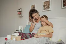 Foto: Mutter sitzt neben ihrem kranken Sohn auf dem Bett und telefoniert mit der Schule.