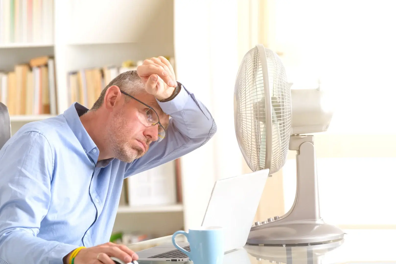 Ein Mann sitzt am Schreibtisch mit Laptop und Ventilator, um sich in der Sommerhitze abzukühlen.