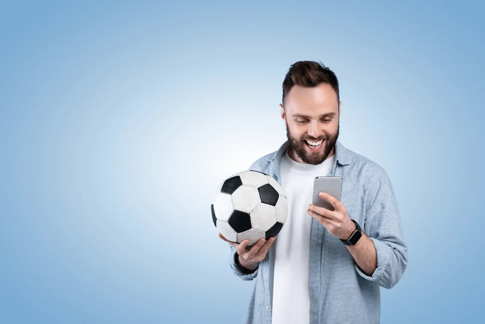 Ein Mann mit einem Fußball in der Hand blickt auf sein Handy und denkt über Sportwetten nach.