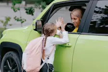 Foto: ein Mädchen, mit Schultasche, verabschiedet sich von ihrer Mutter, die noch im Auto sitzt.