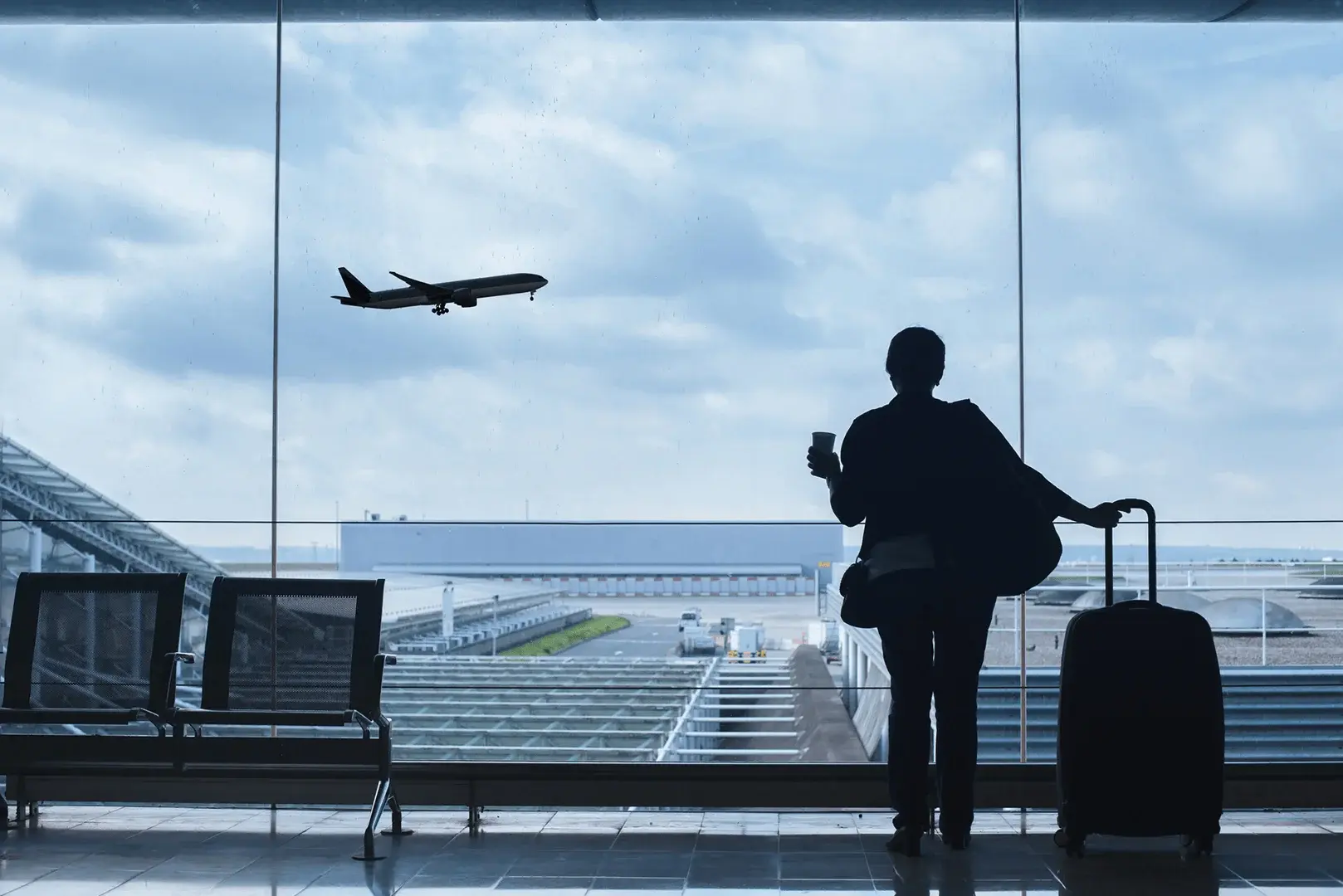 Frau steht mit ihrem Gepäck am Flughafen und sieht ein Flugzeug starten.