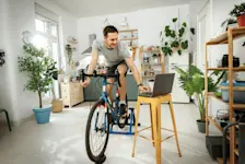 Während der Arbeitszeit fährt ein Mann auf seinem Spinning Bike und arbeitet am Laptop im Homeoffice.