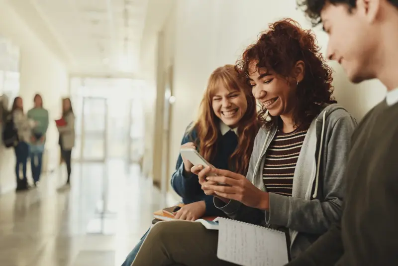 Drei Schüler sitzen auf einer Mauer und schauen auf ihre Handys, während sie über Smartphones im Unterricht diskutieren.