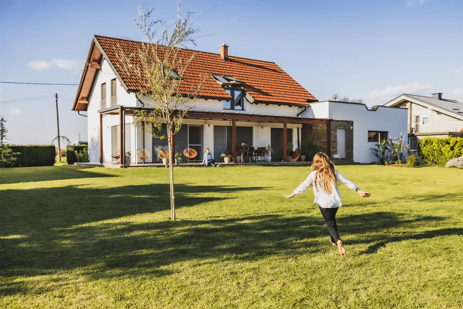 Ein Mädchen läuft spielend durch den Garten,während die Mutter auf der Terrasse sitzt. 