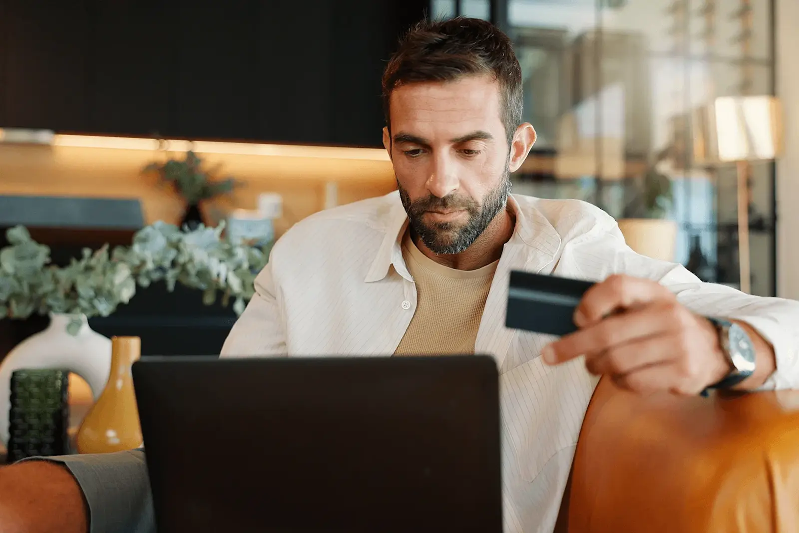 Foto: Ein Mann sitzt mit seinem Laptop in einem Sessel. In einer Hand hält er seine Bankkarte für eine Überweisung.