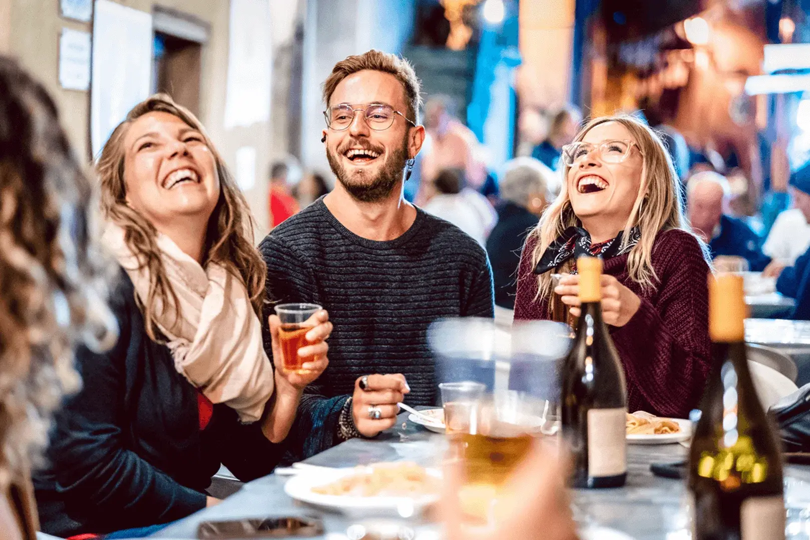Foto: drei Personen sitzen im Außenbereich einer Bar, lachen und trinken Wein.