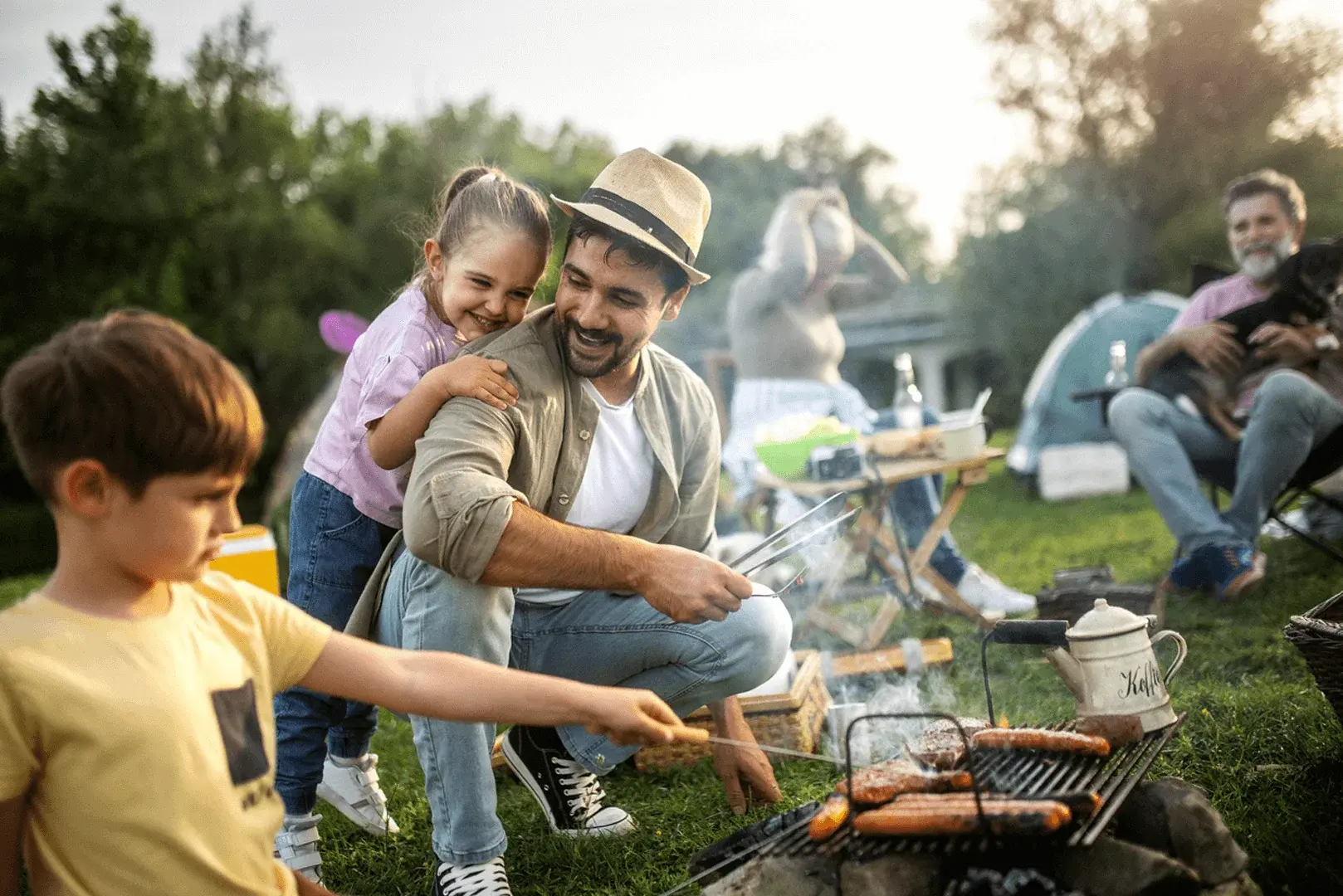 Foto: eine Gruppe von Menschen sitzen im Park. Ein Mann grillt Würstchen mit zwei Kindern.