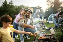 Foto: eine Gruppe von Menschen sitzen im Park. Ein Mann grillt Würstchen mit zwei Kindern.