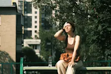 Foto: eine Frau sitzt auf einer Bank in der Stadt, neben ihr steht eine Wasserflasche, sie trocknet ihre Stirn ab.