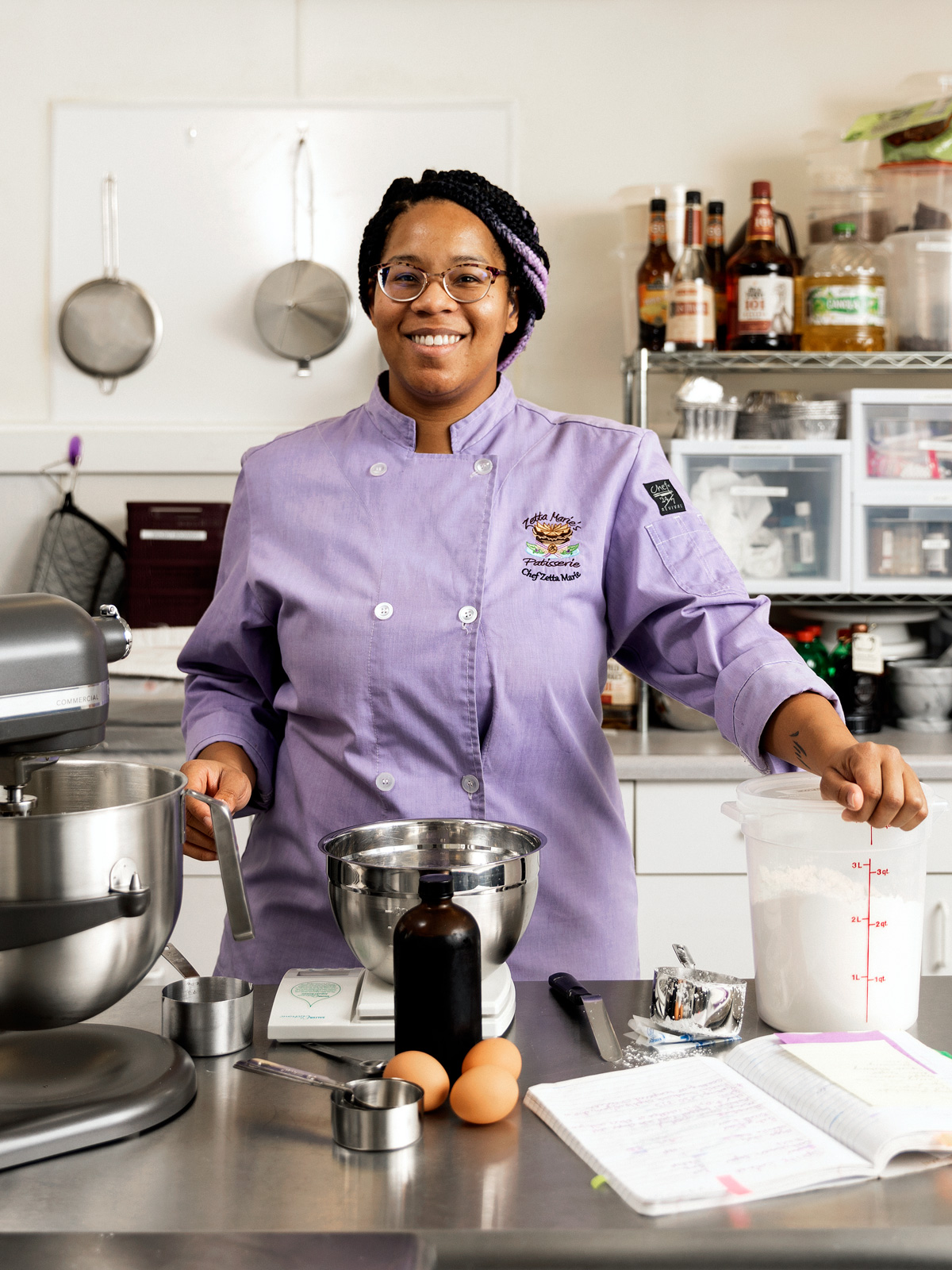 Una mujer con un abrigo de chefs de color púrpura claro en su panadería con la mano tendida en un recipiente de harina

