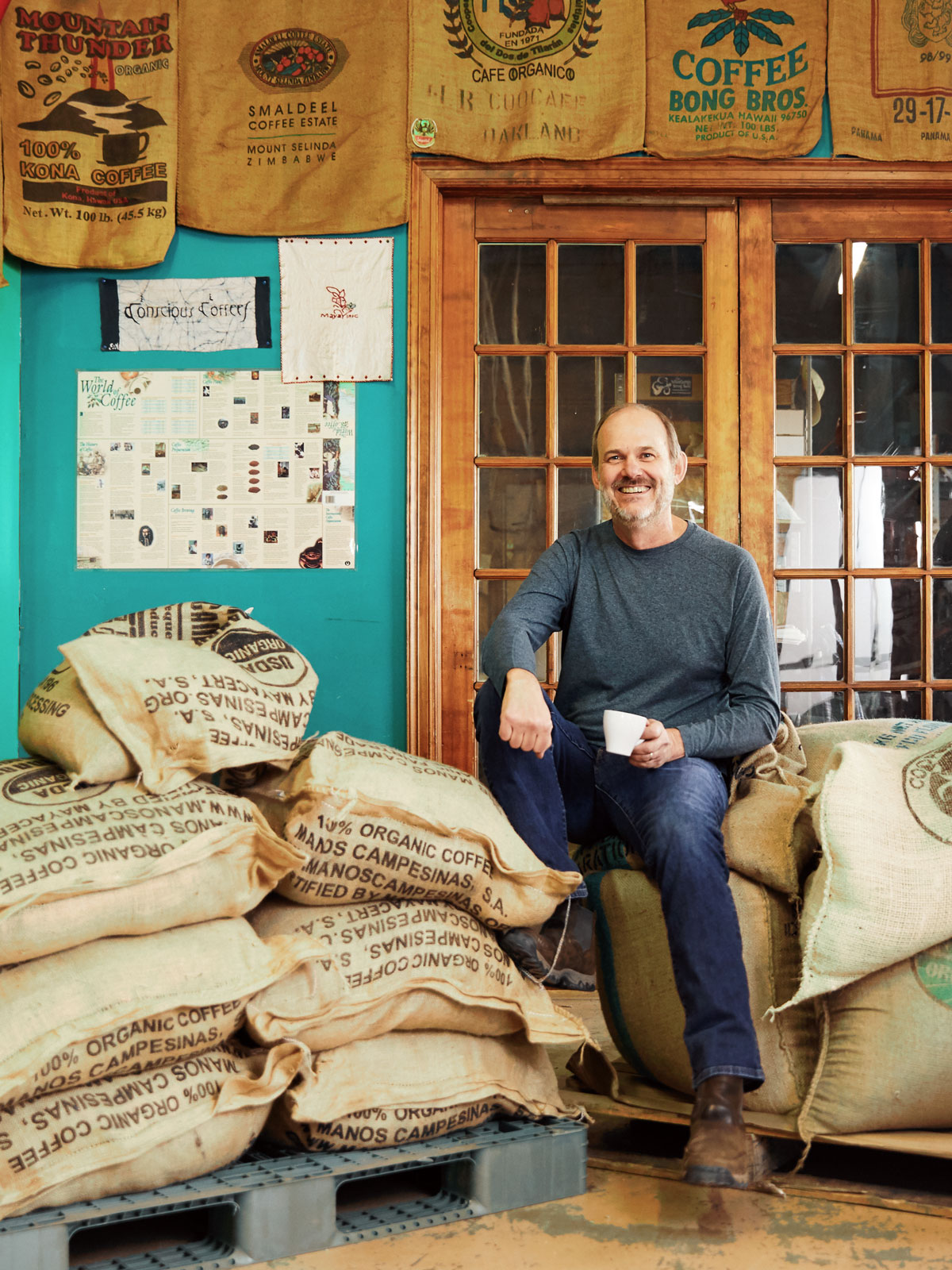 The owner of Conscious Coffees sitting on a stack of burlap bags of coffee beans