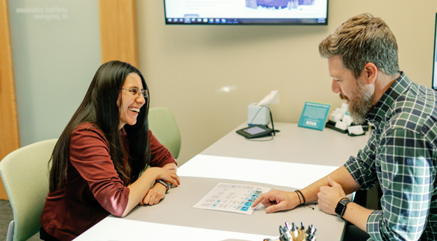 A man and a woman having a discussion across a table.  The man is pointing at a sheet of paper sitting on the table.