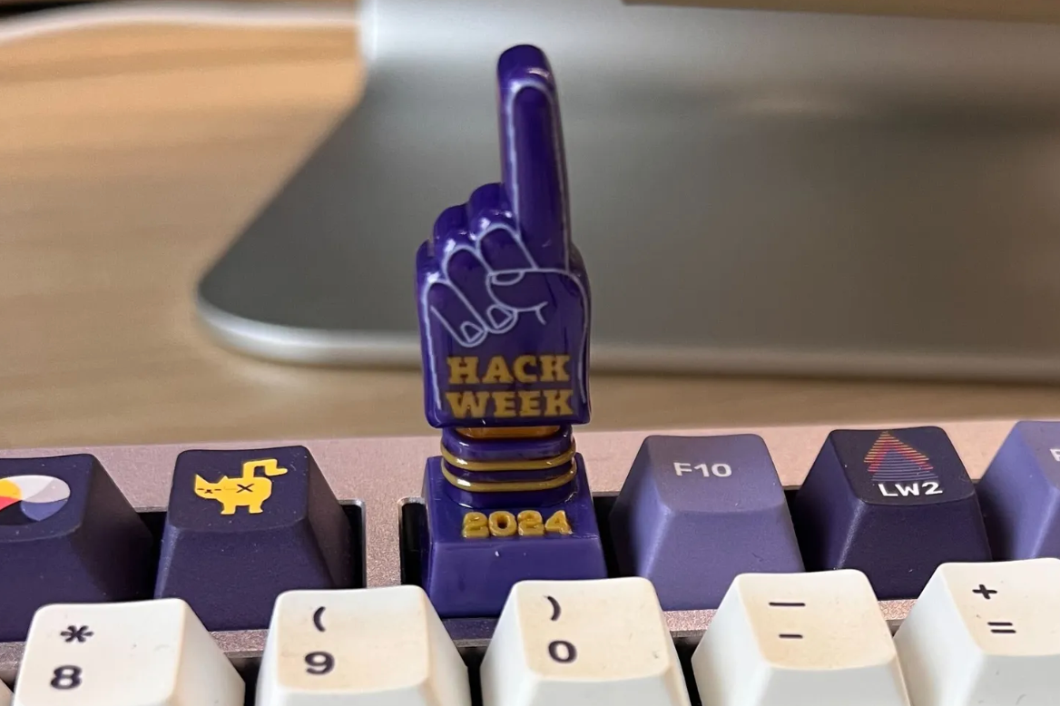 A picture of a keycap for a mechanical keyboard that is on the F9 key. The keycap has a "foam finger" in purple that says "Hack Week 2024"