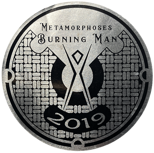 Circular Burning Man sticker labeled “Metamorphoses 2019” featuring the Man symbol and geometric, stone-like pattern in metallic gray.
