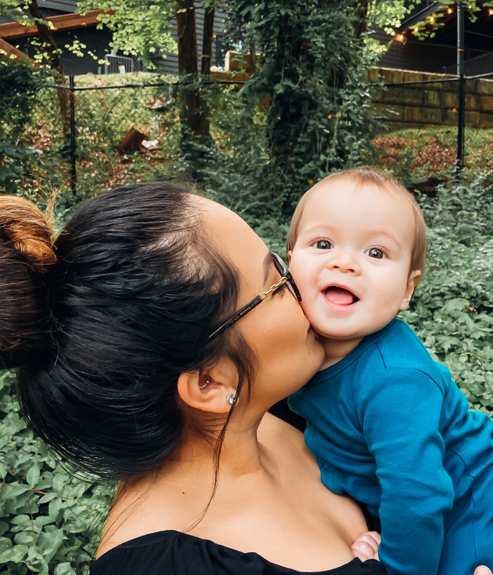 Woman surrounded by green leaves wearing glasses and kissing a baby wearing a teal onesie on the check