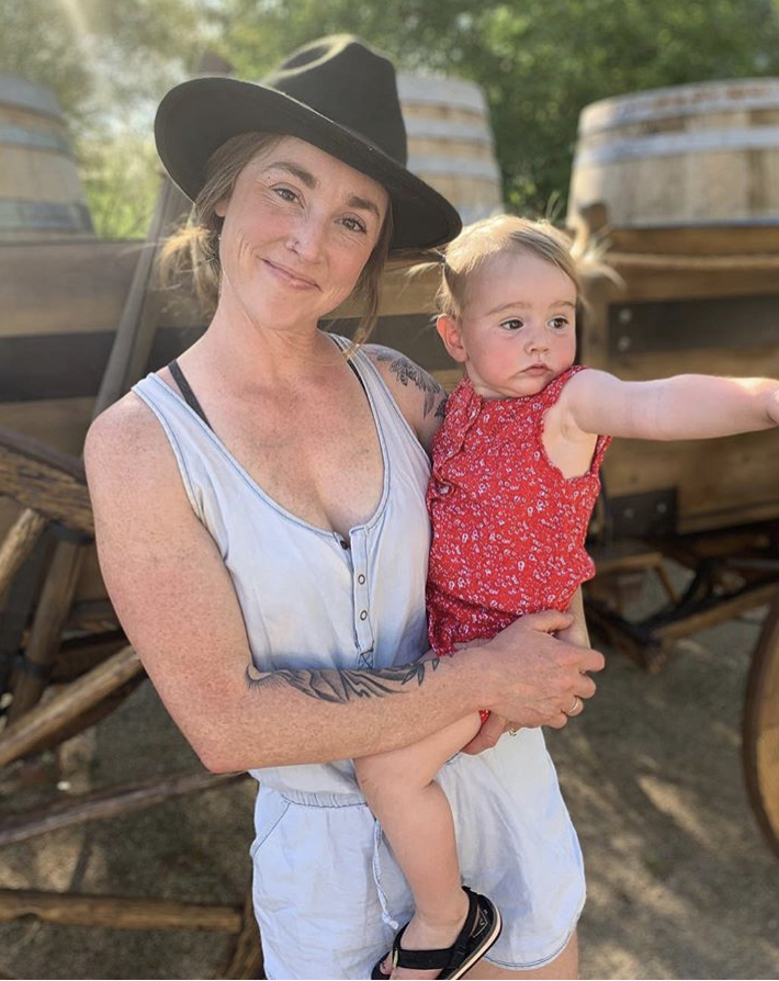 Woman wearing a cowboy hat is holding a little girl who has pigtails and is wearing a pink summer outfit