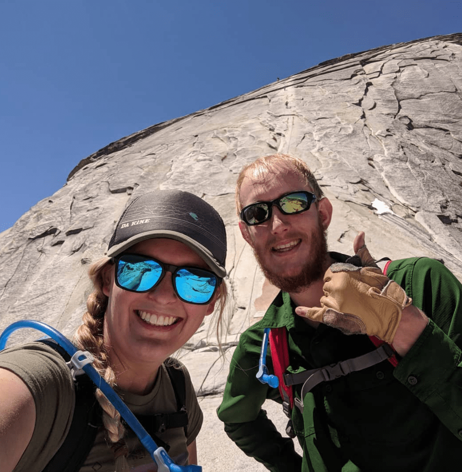 Woman and a man smiling wearing rock climbing gear standing on the side of a mountain
