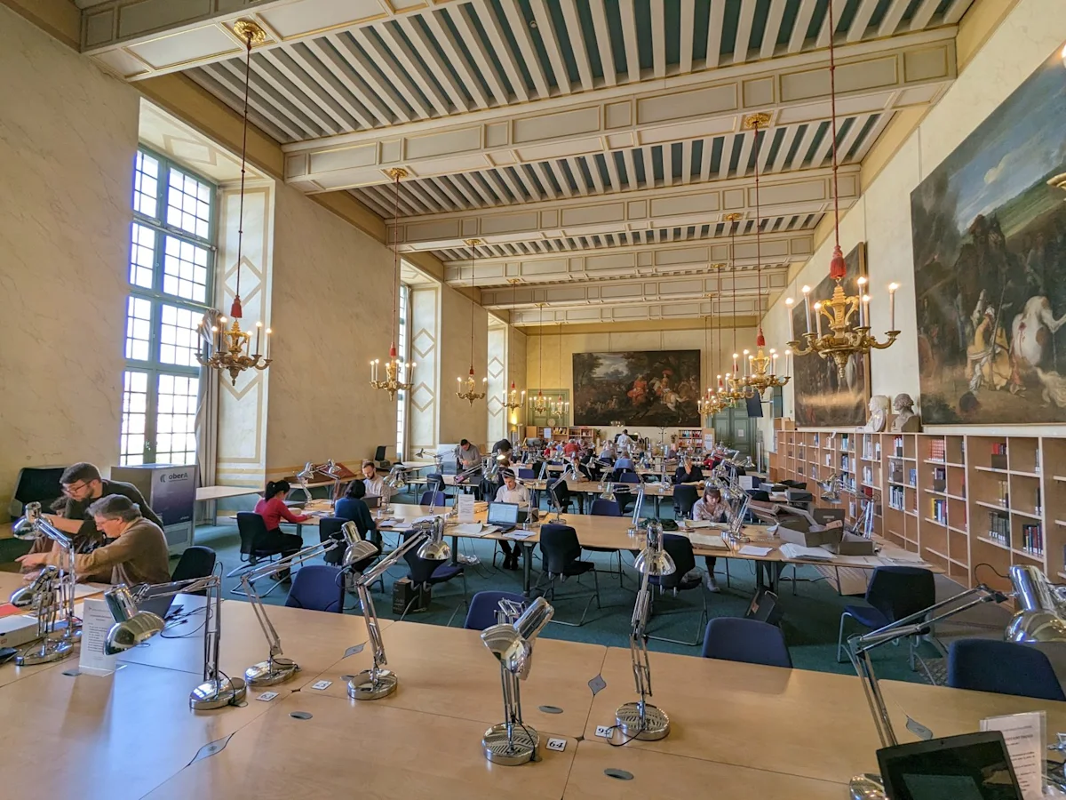 Readers at long tables in a high reading room with chandeliers and large paintings on the walls