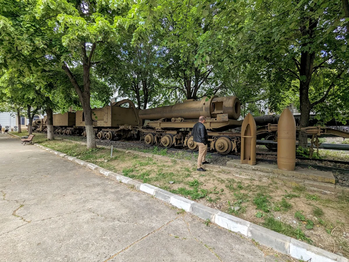 A line of rail wagons under trees carrying the barrel and parts of a siege howitzer, with two large shells standing beside them and a visitor for scale