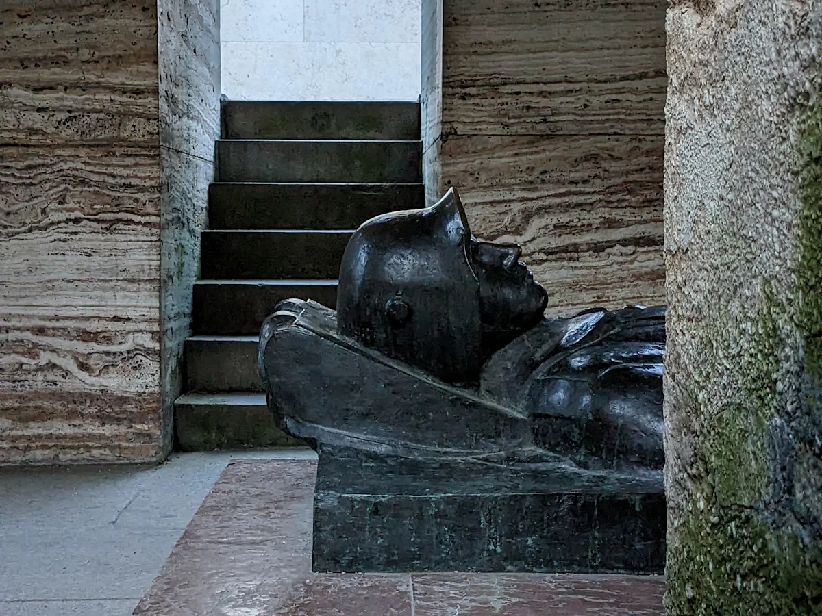 Bronze figure of a dead soldier on a tomb, seen in profile between the stone pillars of the crypt of the Munich war memorial