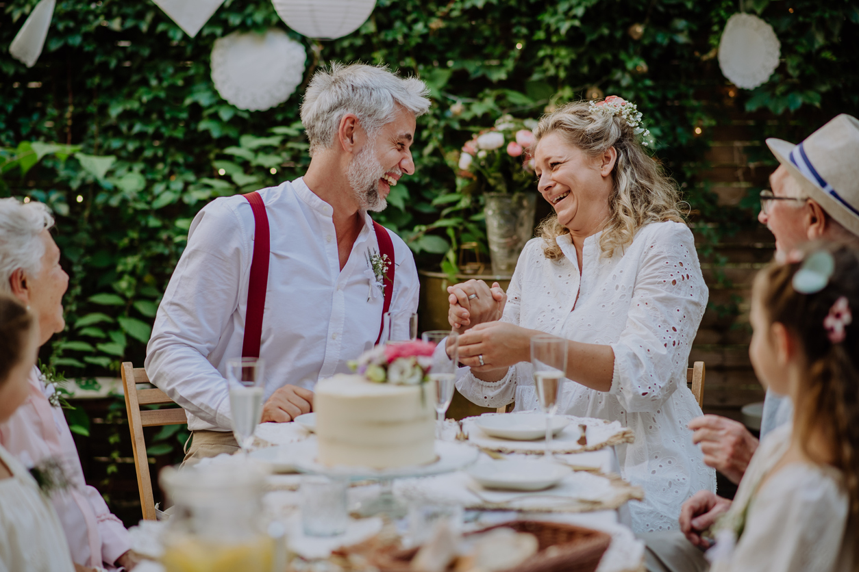 An elderly couple joyfully celebrates an anniversary at a table set with a cake and flowers. They are surrounded by family in a lush garden setting.