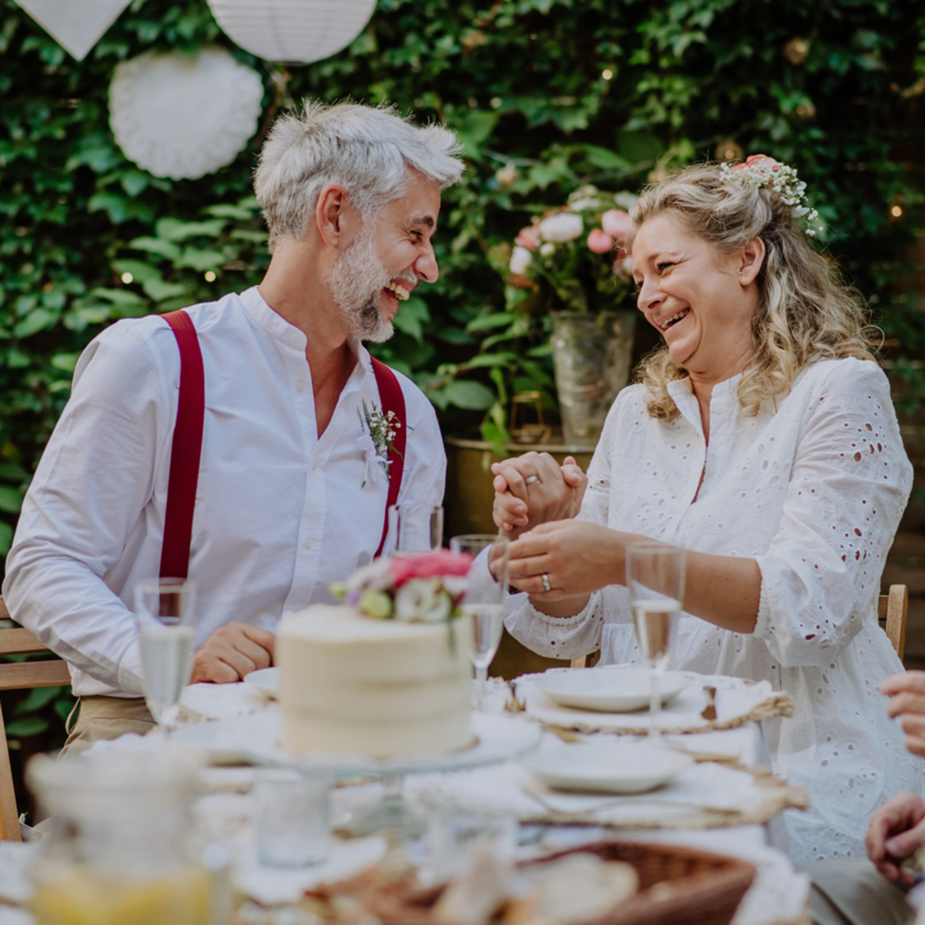 An elderly couple joyfully celebrates an anniversary at a table set with a cake and flowers. They are surrounded by family in a lush garden setting.