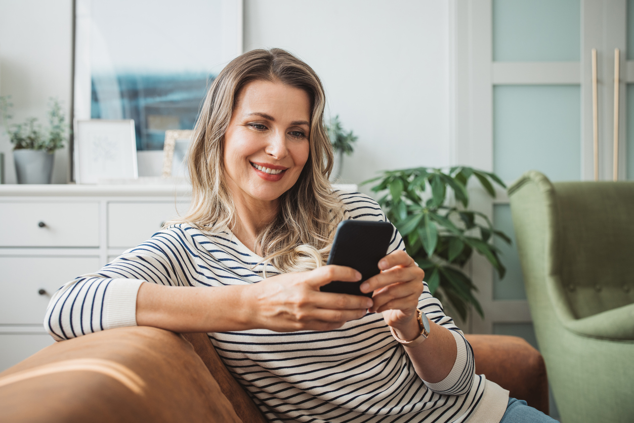 Smiling woman in striped shirt sits on a couch, looking at her smartphone. The room is cozy, with plants and framed art, creating a warm, relaxed atmosphere.